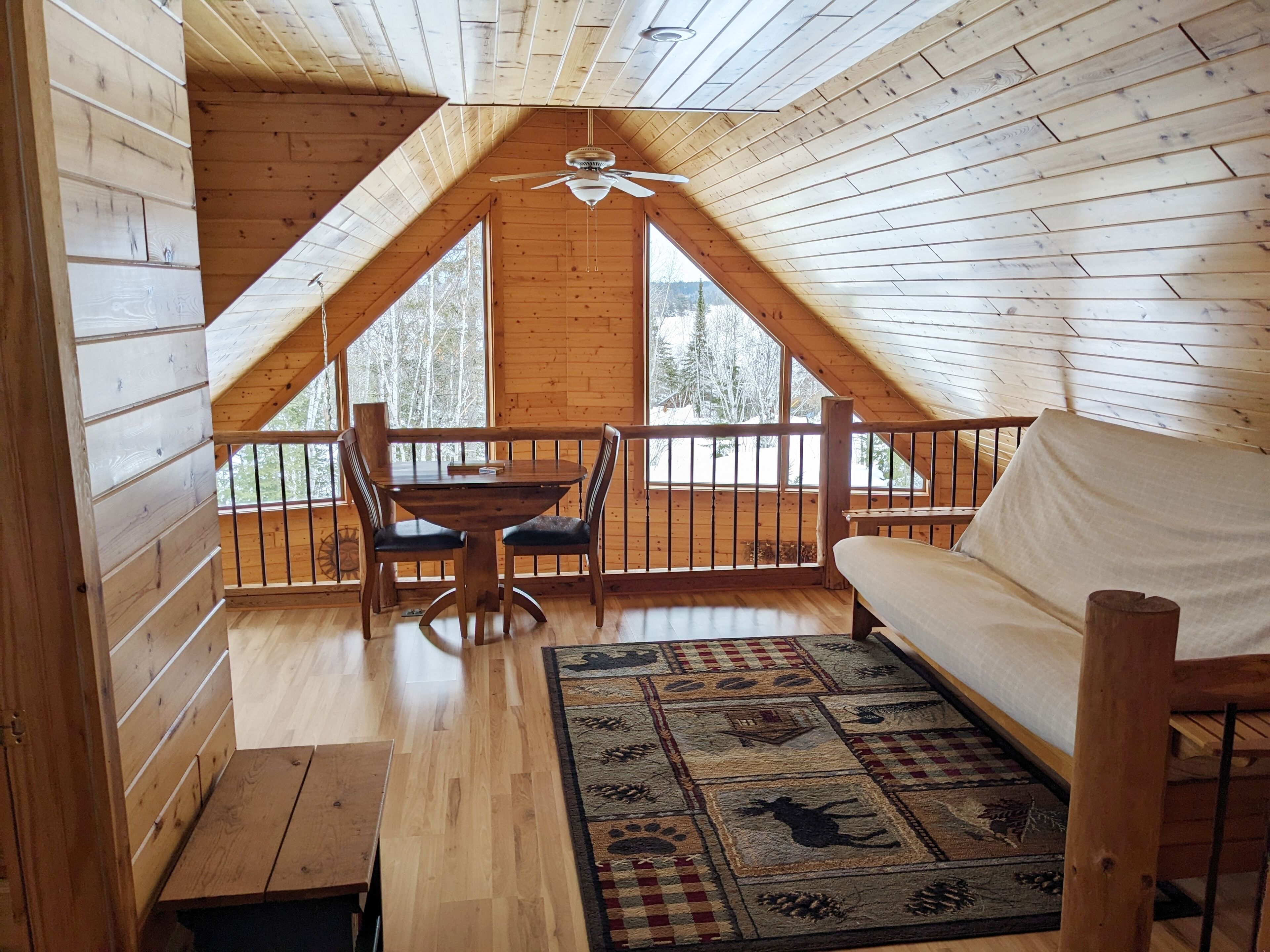 A cozy cabin with a wooden interior, a dining area with a table and chairs, a sofa, and a large window offering a view of trees.