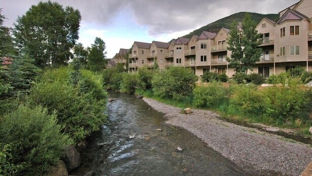 A row of modern multi-story buildings with balconies, a river in the foreground, and a hilly landscape in the background. - Ski Trip Value! Walk Everywhere, Hot Tub, Top Floor 1 Bedroom Condo by RedAwning (Telluride)