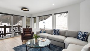 Uma sala de estar moderna com uma poltrona de couro, um sofá cinzento e uma mesa de centro de vidro. Há vista para uma paisagem nevada através de grandes janelas.