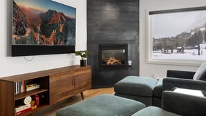 Uma sala de estar moderna com lareira, uma grande pintura de paisagem de montanha e vista para uma paisagem nevada lá fora.