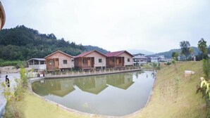 A row of wooden cabins with a central pond, surrounded by greenery and hills.