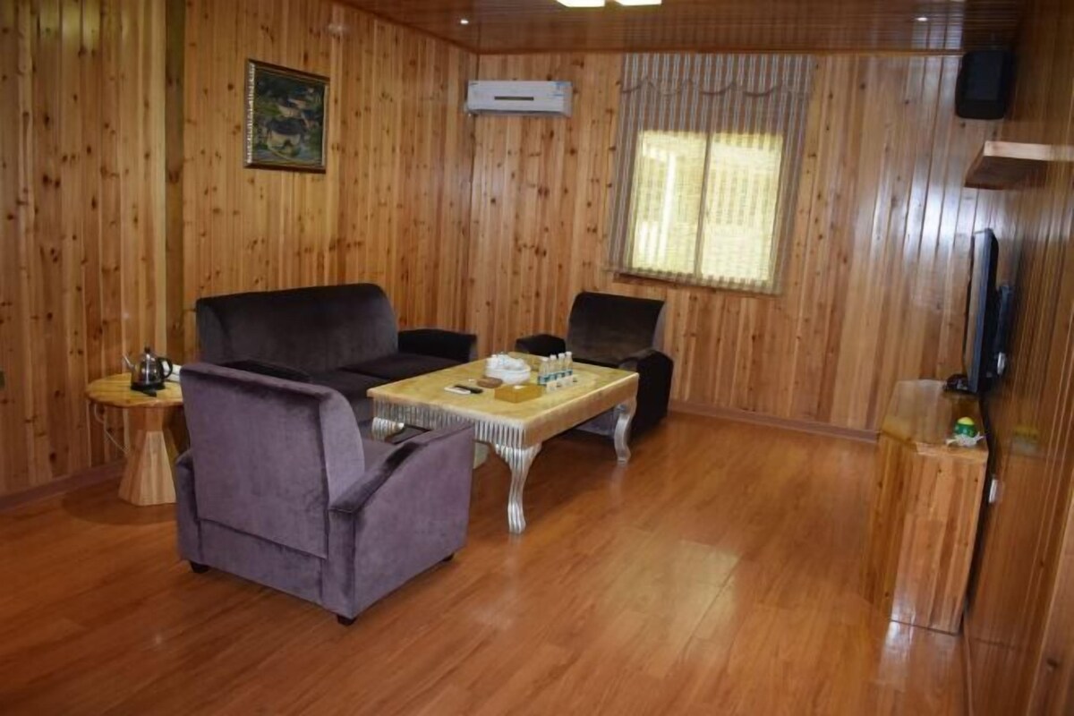 A wooden-paneled room with a sofa, two armchairs, a small table, a television, and a window with curtains.