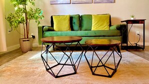 Un canapé vert avec des coussins jaunes, deux tables hexagonales, une plante en pot et un tableau accroché au mur.