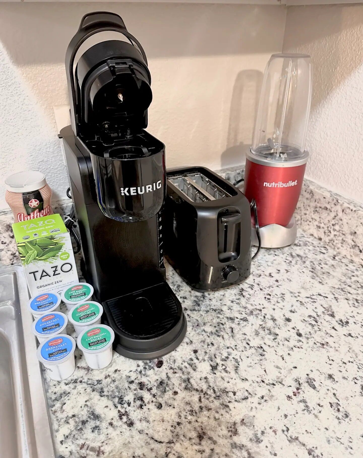 Eine Küchenarbeitsfläche mit einer Keurig-Kaffeemaschine, einem Toaster, einem Nutribullet-Mixer und mehreren Behältern mit Tazo Bio-Kaffee.