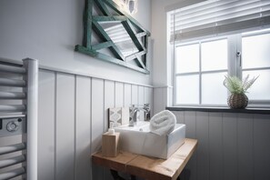Un baño con un lavamanos blanco, un estante de madera con artículos de aseo, una toalla y una ventana con persianas.