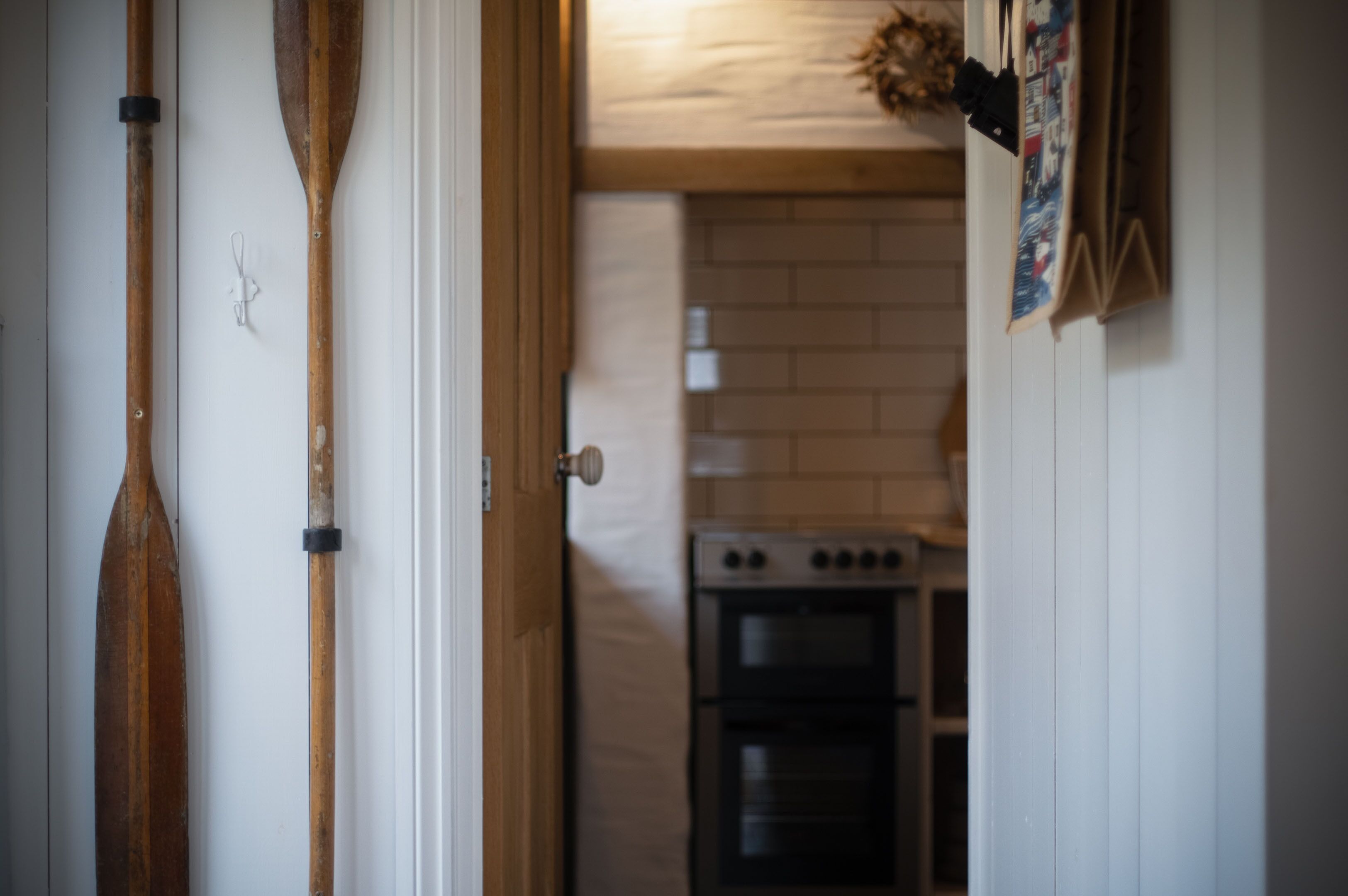 A narrow hallway with wooden paddles leaning against the wall, a door, and a kitchen area with a stove and oven in the background.