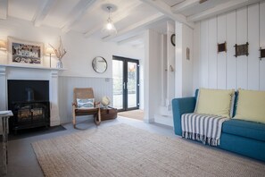 A living room with a fireplace, a blue sofa, a wicker chair, and a large window. - The Old Spa - 2 Bedroom Cottage - Pleasant Valley (Narberth)