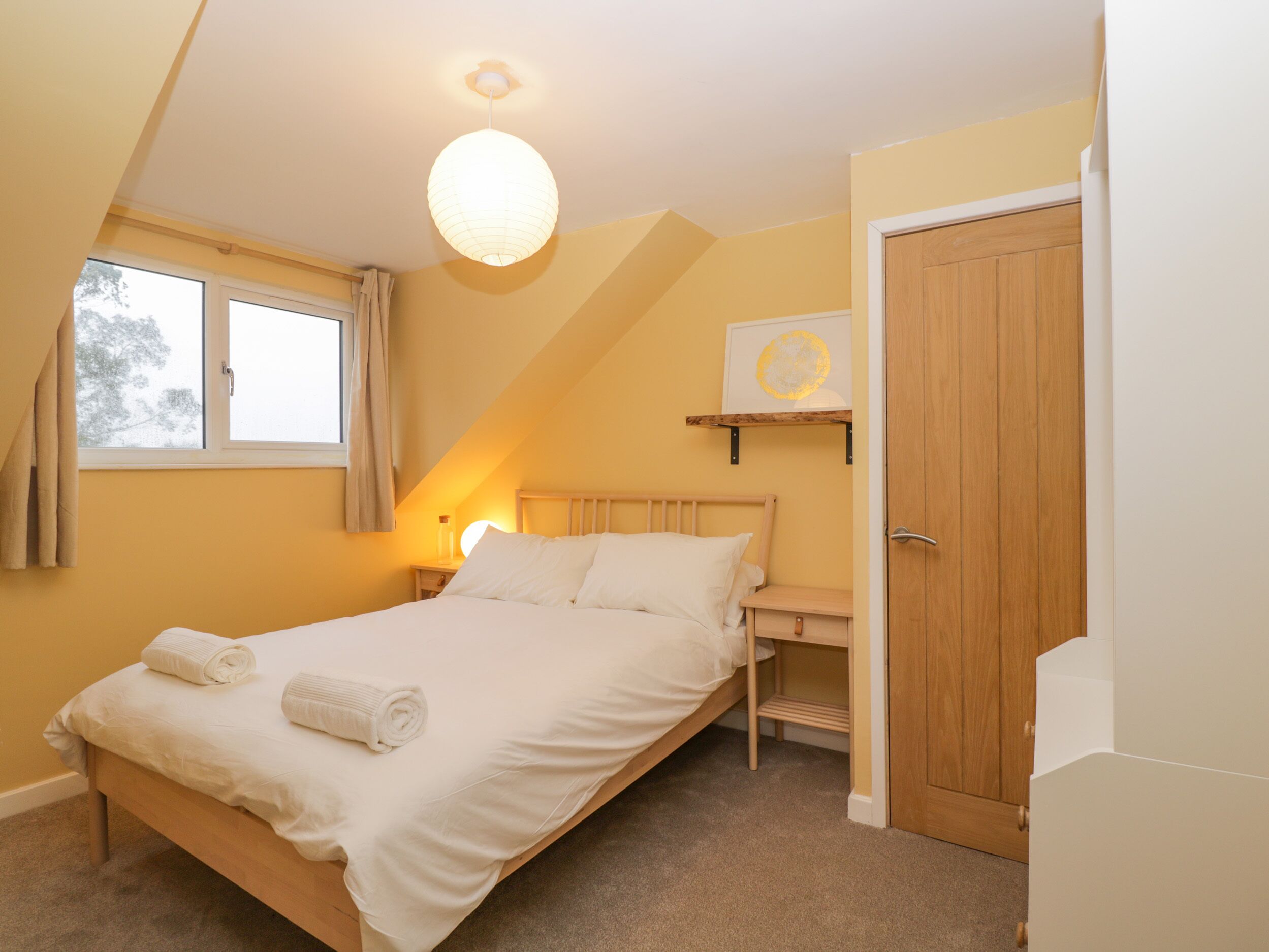 A bedroom with a bed, a bedside table, a lamp, a window, and a door.