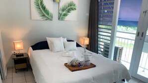 A neatly made bed with white linens and a dark headboard, two bedside lamps, a tray with toiletries, and a view of the outdoors through a large sliding glass door.