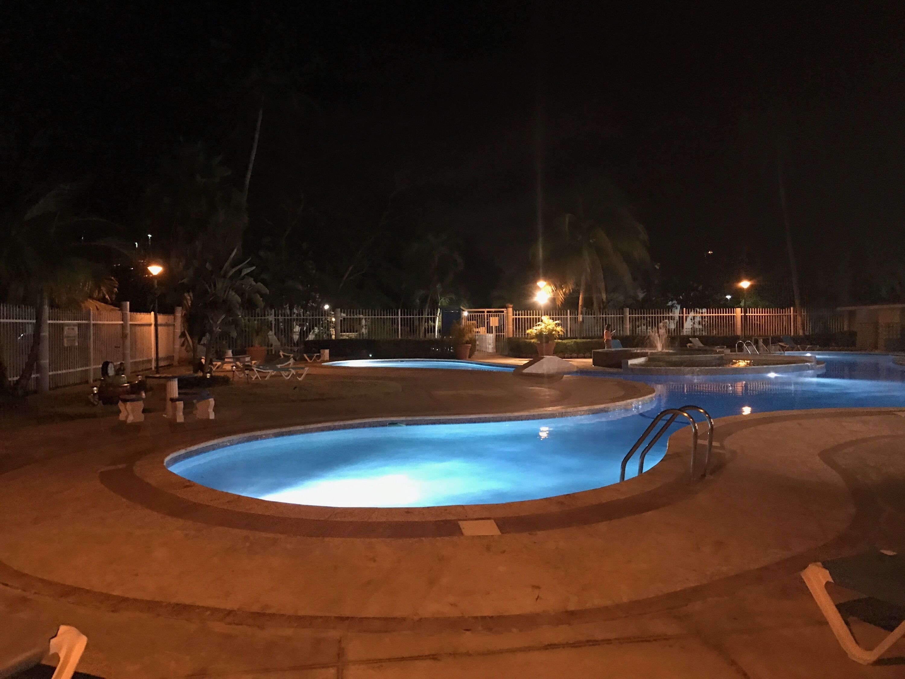 Un espace piscine nocturne comprenant plusieurs piscines, des chaises longues et un périmètre délimité par une clôture.