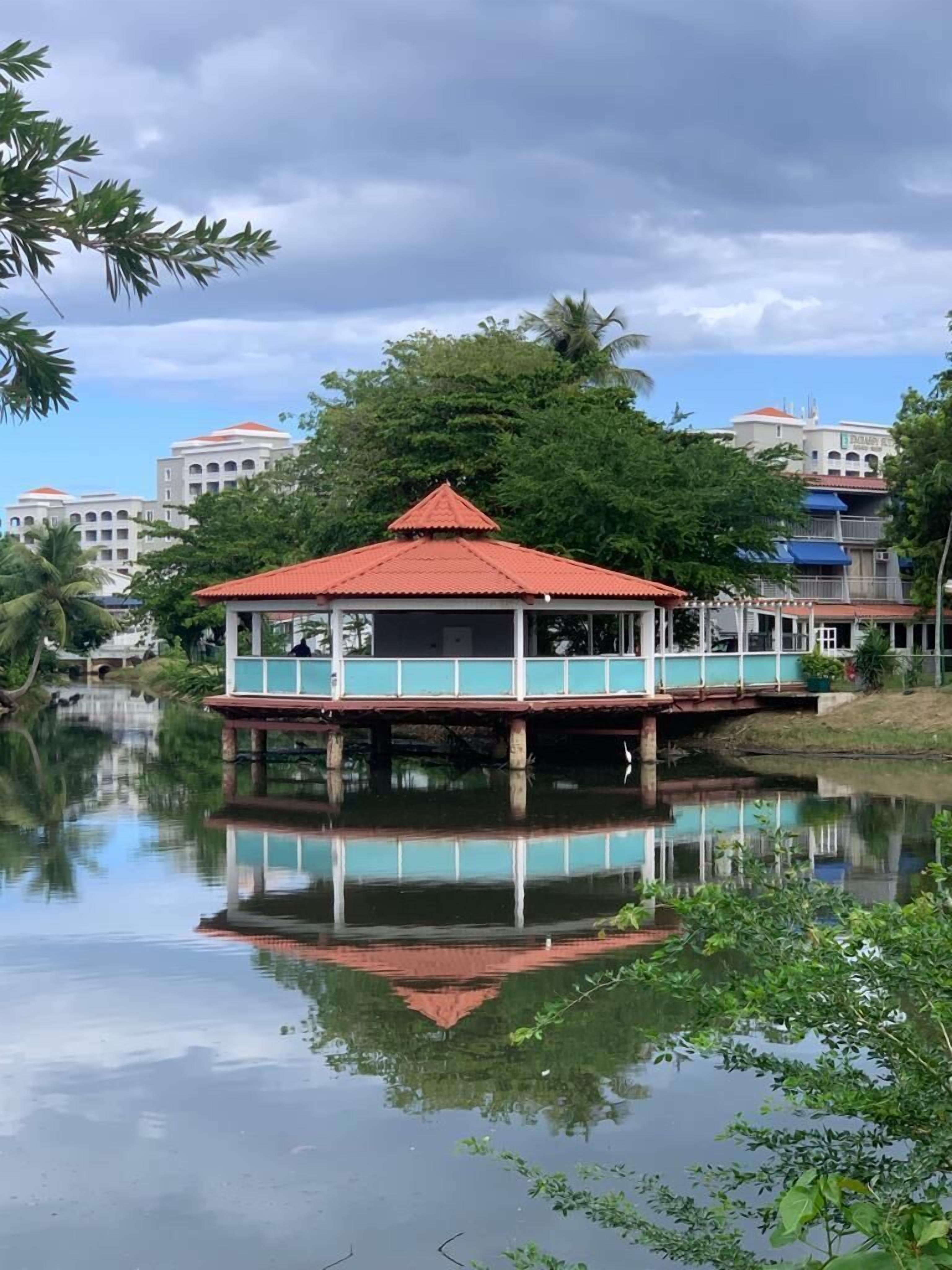 A gazebo-like structure with a red roof, situated on stilts over a calm body of water, surrounded by lush greenery and modern buildings in the background.
