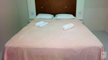 Una cama individual con cabecera de madera, dos almohadas y dos toallas enrolladas sobre una colcha rosa.