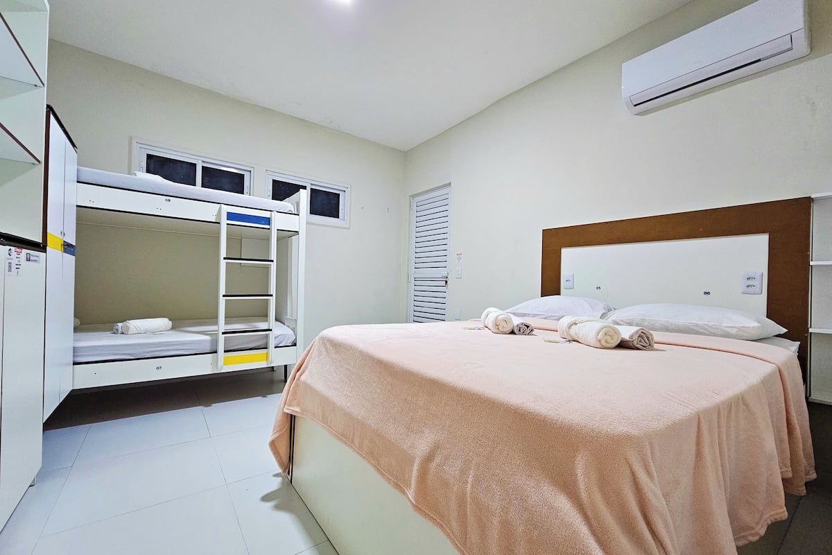 Una habitación de hotel con literas, una cama grande con una manta rosa y un equipo de aire acondicionado en la pared.