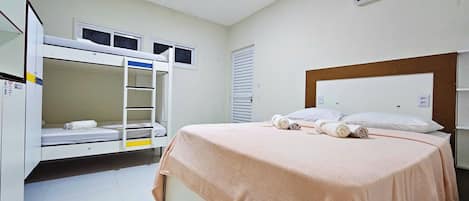 A hotel room with bunk beds, a single bed with a pink blanket, an air conditioning unit, and a window with blinds.