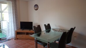 A dining area with a glass table, black chairs, a television on a wooden stand, and a clock on the wall.