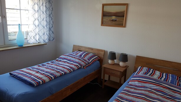 A bedroom with two single beds, a window with curtains, a small table with two lamps, and a painting on the wall. - Dornum" vacation apartment (Dornumersiel)