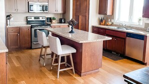 A modern kitchen with wooden cabinets, stainless steel appliances, a central island with bar stools, pendant lighting, and a large window providing natural light.