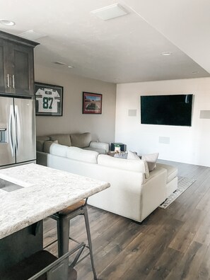 A modern kitchen with a marble countertop and bar stools, a white sofa set, and a flat-screen TV mounted on the wall.