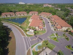 Vista aérea de una zona residencial con varias construcciones de dos pisos, una carretera curva y un lago pequeño con fuentes.