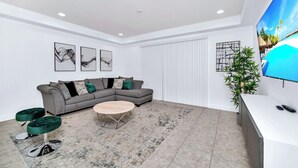 A modern living room with a grey sectional sofa, a round coffee table, and a large flat-screen TV mounted on the wall. - Big Screen Movie Nights! Champions Gate Resort Close 2 Disney! 8165 (Davenport)
