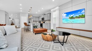A modern living room with a sofa, ottoman, and coffee table, a dining area with chairs, a kitchen with cabinets and appliances, and a large TV on the wall displaying a beach scene.