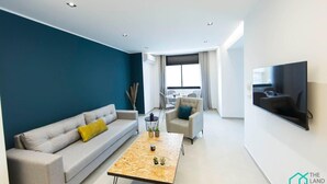 A modern living room with a sofa, armchair, and coffee table, a large window with curtains, and a mounted TV. - Sunset 202 Luxury With gym Club in Lac 1 (Tunis)