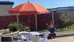 Une terrasse avec une table ronde et quatre chaises blanches, un parasol orange et une plante en pot.