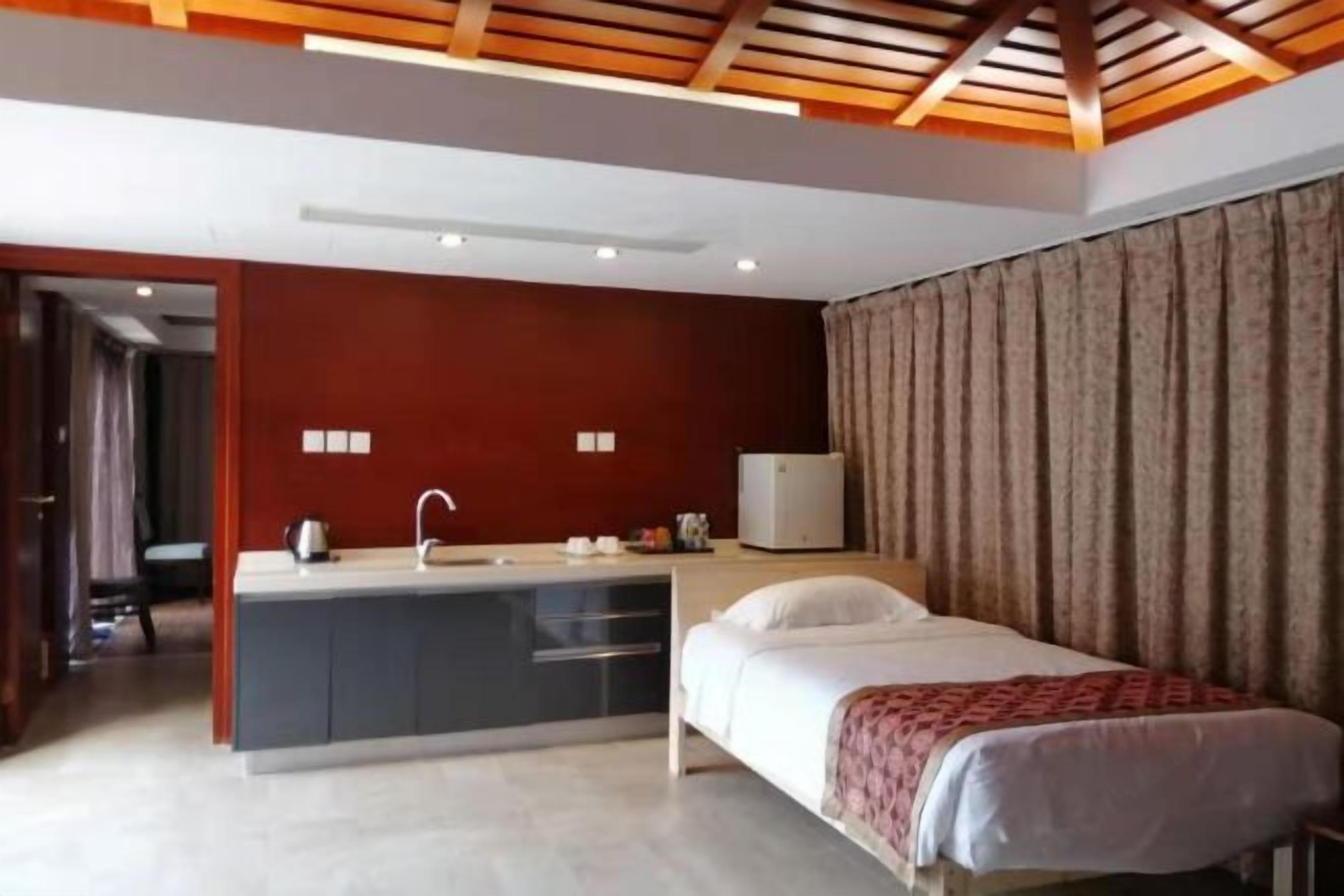 Un dormitorio con cama, una mesita de noche, escritorio con silla, ventana con cortinas y una kitchenette con lavabo y refrigerador.
