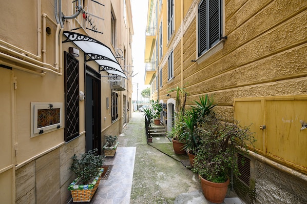 A narrow alleyway with buildings on either side, potted plants, and a small staircase leading to a door.