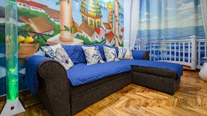 A cozy room with a blue sofa, a mural of a coastal scene, and a view of the sea through a window.