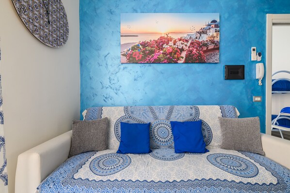 A room with a bed, a blue wall, a painting of a coastal scene, and a phone on the wall.