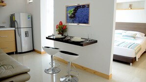 Una habitación de hotel compacta con kitchenette, cama con ropa de cama a rayas, un pequeño comedor con una mesa y dos taburetes altos, y un televisor montado en la pared.