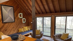 L’intérieur chaleureux d’un chalet avec un plafond en bois, un canapé bleu, un fauteuil marron et une grande fenêtre donnant sur le paysage extérieur.