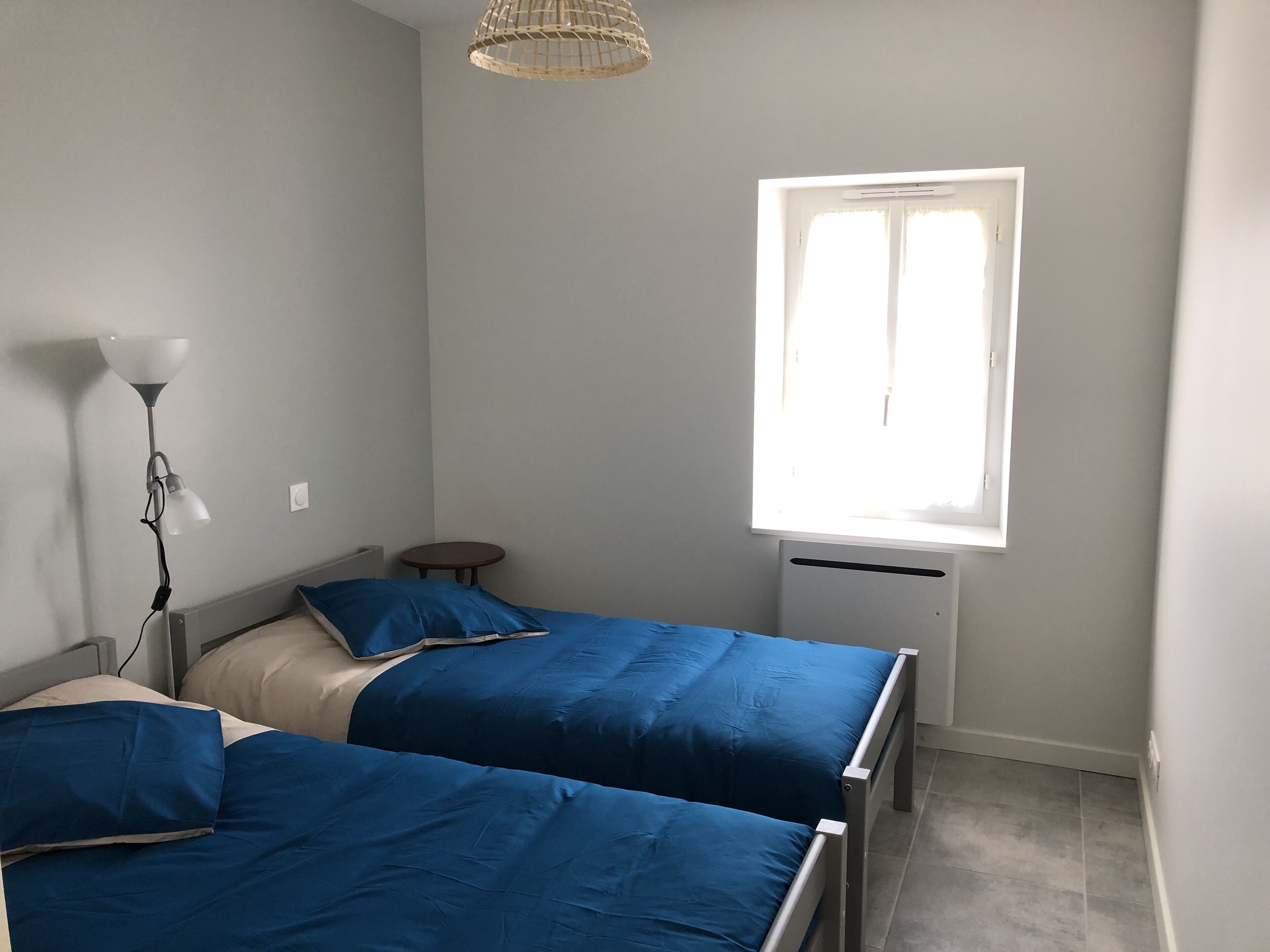 Een slaapkamer met twee bedden, een klein rond tafeltje, een vloerlamp en een raam met jaloezieën.