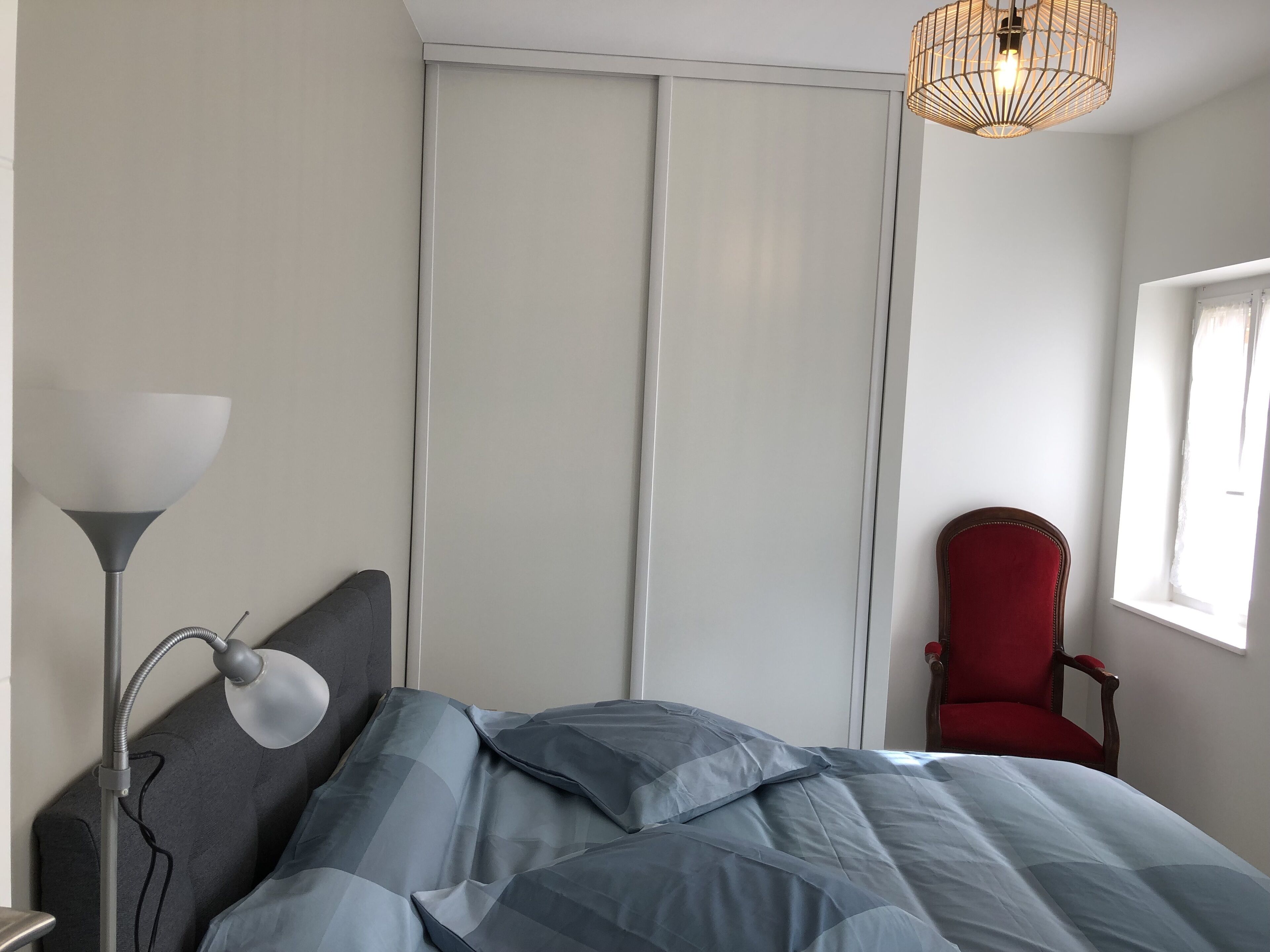 Ein Schlafzimmer mit einem Bett, einer Schreibtischlampe, einem roten Sessel, einem Kleiderschrank und einem Fenster.