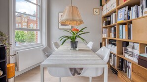Un comedor con una mesa, sillas, una lámpara colgante, una planta en maceta y una estantería llena de libros y carpetas.