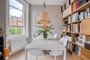 A dining area with a table, chairs, a hanging lamp, a potted plant, and a bookshelf filled with books and binders.