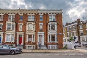 A row of red brick townhouses with bay windows and white trim, a red door, and a street view with cars.