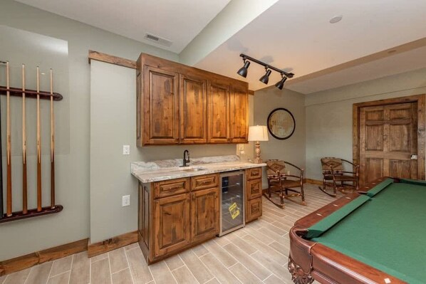 A pool table with a green felt surface, wooden cabinets with a marble countertop, and a wall-mounted lamp.