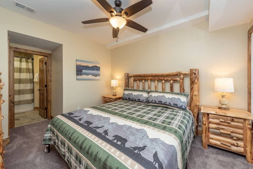A bedroom with a bed, wooden headboard, two bedside tables with lamps, a ceiling fan, and a door leading to another room.