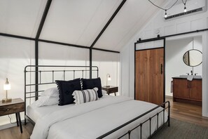 A modern bedroom with a metal bed frame, white bedding, a wooden wardrobe, and a small bedside table with a lamp. - Cherry Cove Glamping W/ Hot tub AND Cowboy Pool! (Fredericksburg)