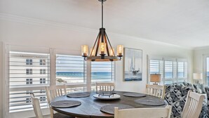 Un espace repas avec une table ronde, des chaises blanches, un lustre et une vue sur l’océan grâce à de grandes fenêtres.