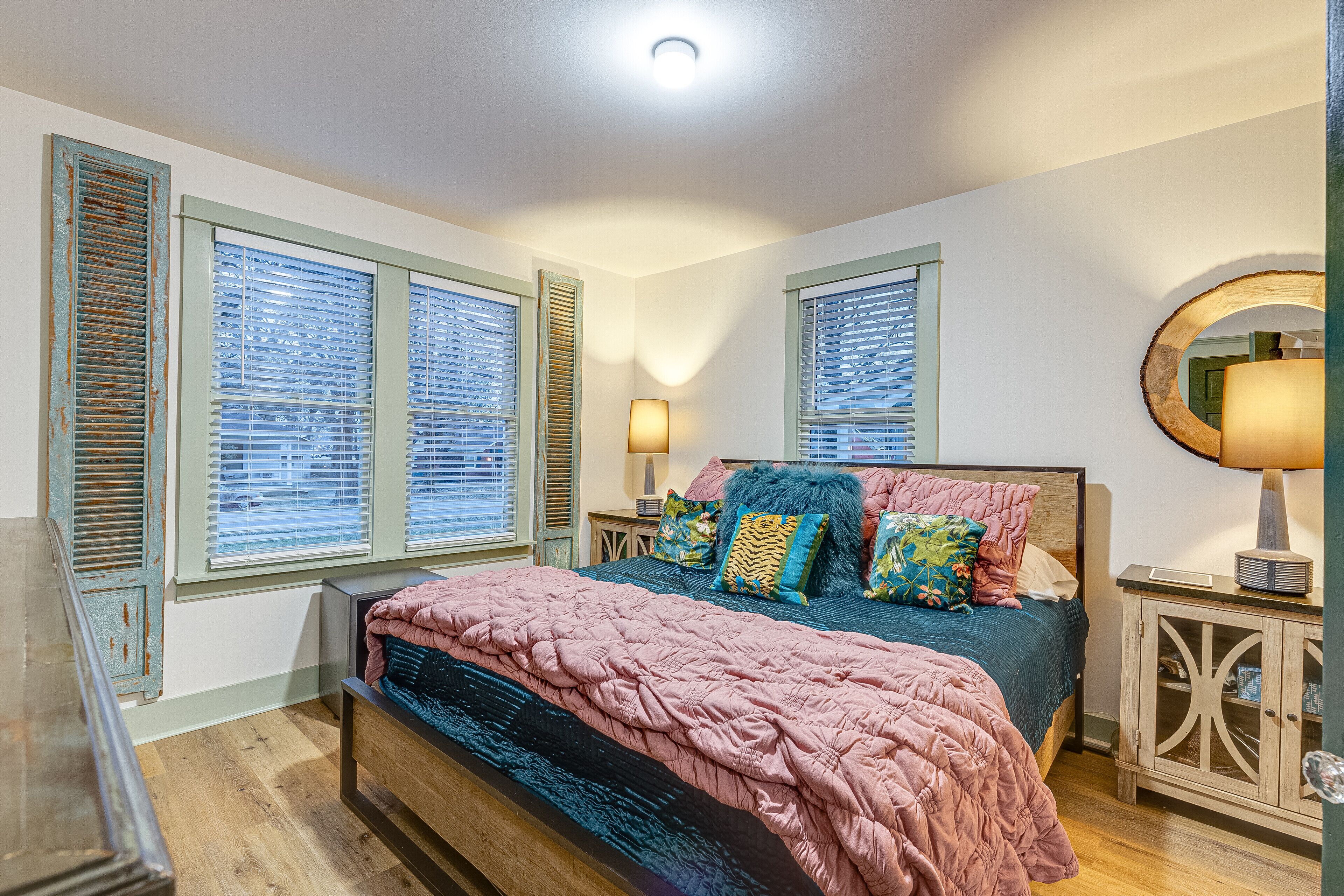 A bedroom with a bed, two bedside tables, a mirror, and a window with shutters.