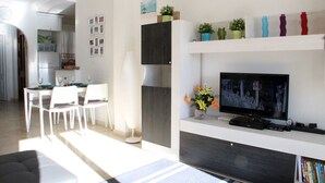 A modern living room with a flat-screen TV, a dining area with white chairs, and a kitchen in the background. - 2 Bed 2 Bath Beautiful Apt Next to Villamartin Plaza (Orihuela Costa)