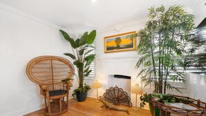 A room with a wooden floor, a rattan chair, a large potted plant, a decorative wall piece, and a fireplace.