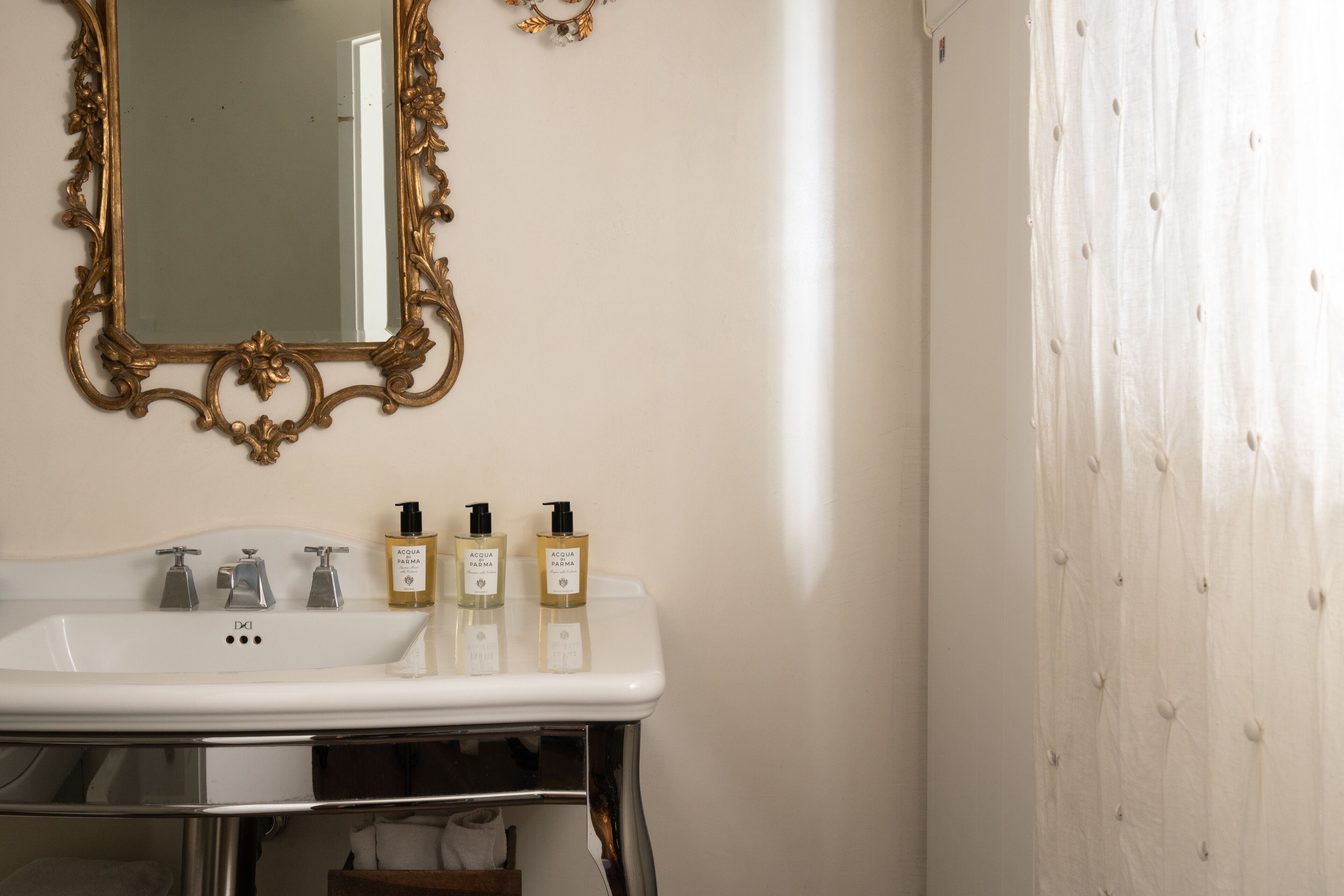 A bathroom with a white sink, gold ornate mirror, and three bottles of Acqua Paloma on the counter.