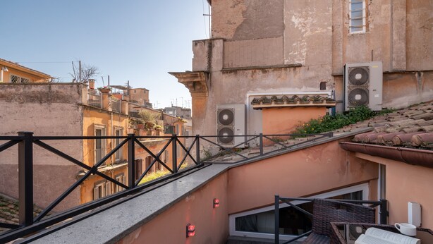 Een smalle trap die leidt naar een balkon met een rieten stoel en een tafel, omringd door gebouwen met balkons en airconditioners.