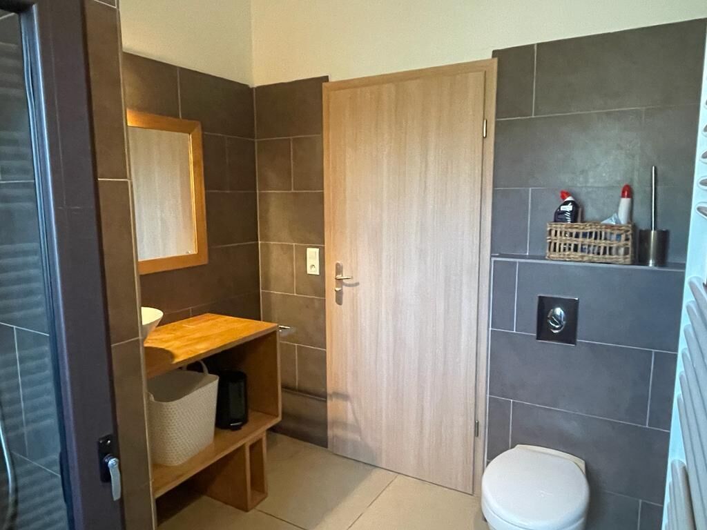 Ein Badezimmer mit Holztür, Toilette in Weiß, Ablage für Toilettenartikel und einem Spiegel.