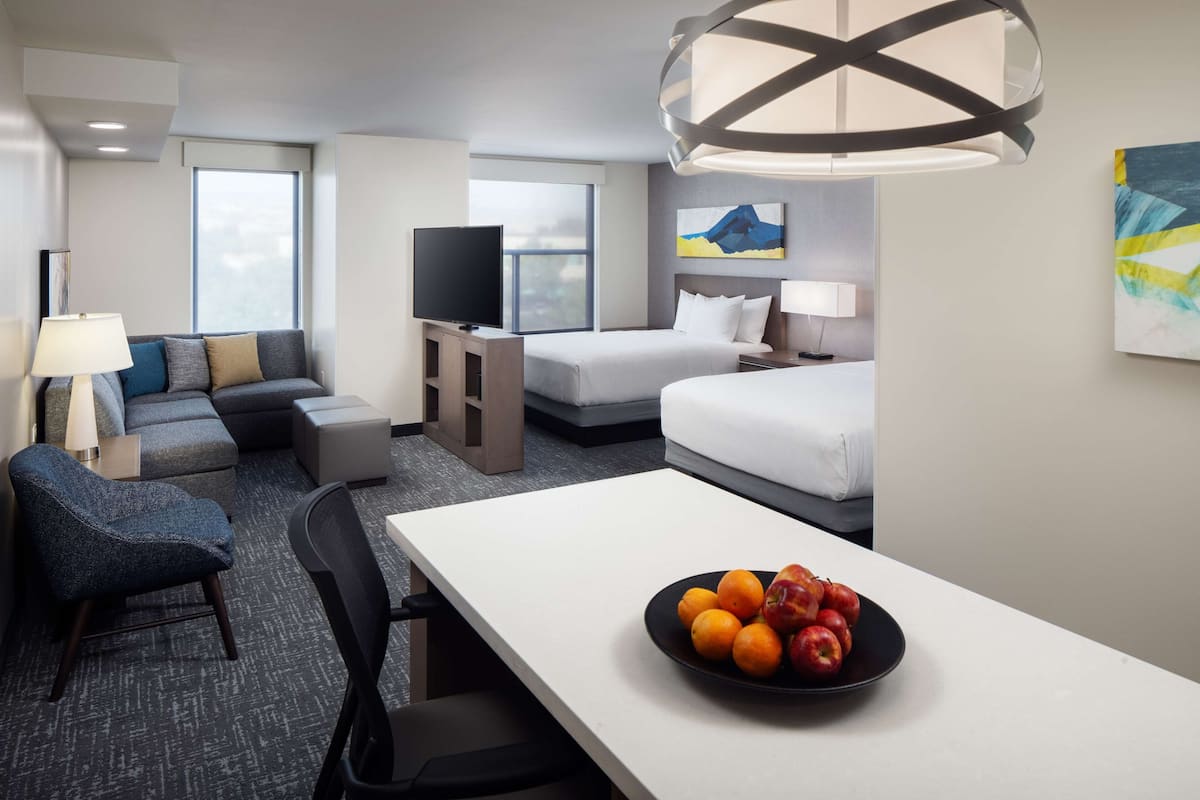 Hotelkamer met een bank, twee bedden, een televisie, een eettafel met fruit en kunstwerken aan de muur.