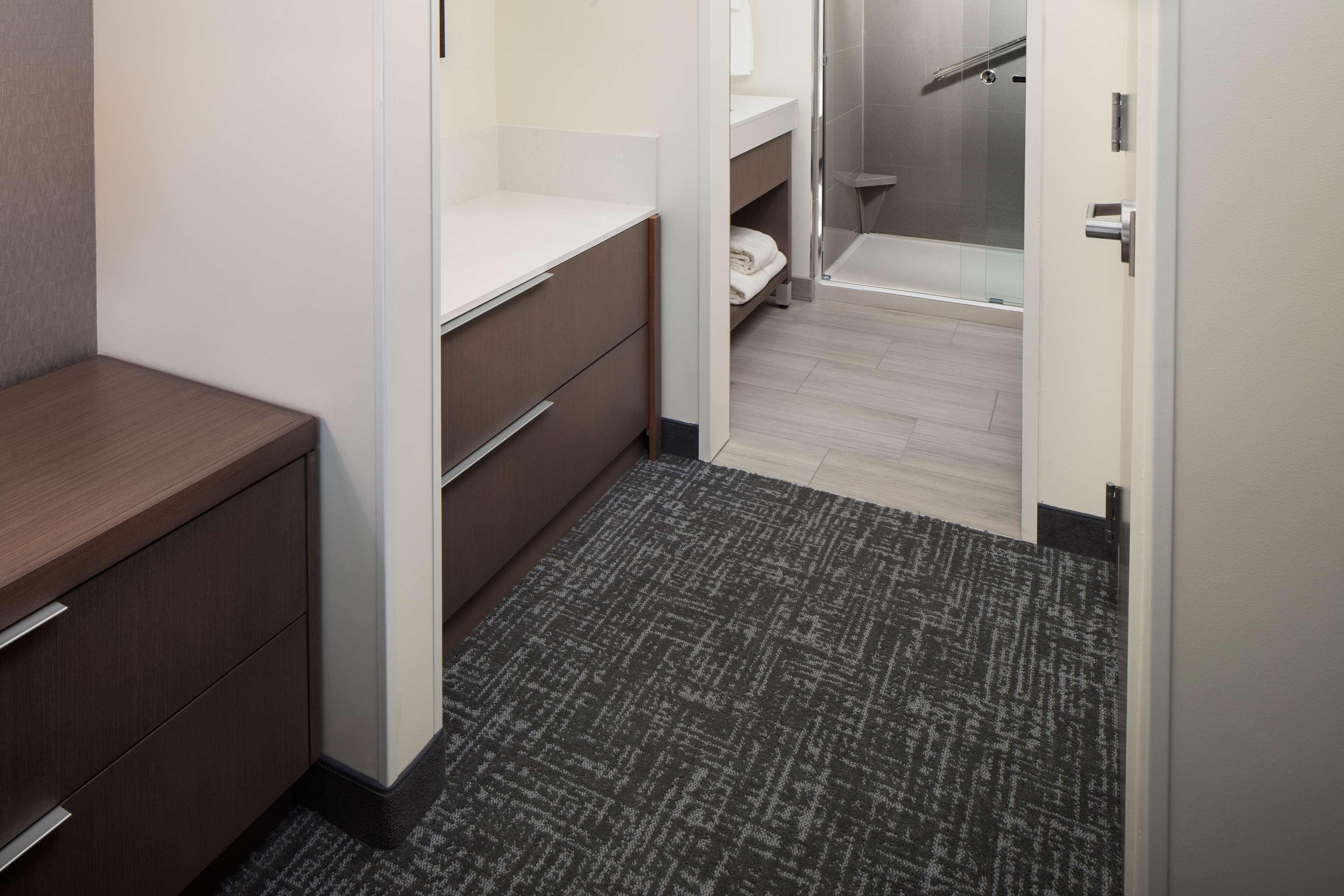 A hotel room with a wooden desk, a white vanity, a shower area, and a carpeted floor.